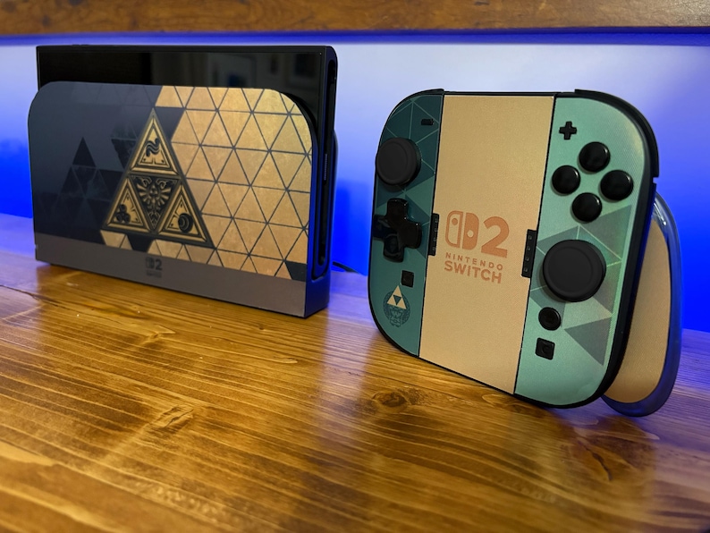 May include: A Nintendo Switch console dock and a Joy-Con controller grip featuring a custom skin with a geometric pattern in shades of teal, tan, and dark blue. The design includes a central triangular emblem and the text Nintendo Switch 2.