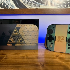 May include: A Nintendo Switch console dock and a pair of Joy-Con controllers featuring a gold and dark green geometric pattern. The dock displays a central triangular emblem, while the controllers show the Nintendo Switch 2 logo in black text.