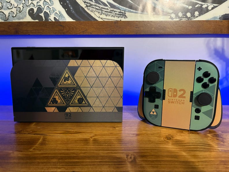 May include: A Nintendo Switch console dock and a pair of Joy-Con controllers featuring a gold and dark green geometric pattern. The dock displays a central triangular emblem, while the controllers show the Nintendo Switch 2 logo in black text.