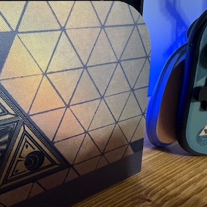 May include: A gaming console dock and a handheld controller featuring a blue and gold geometric triangle pattern. The controller displays a small gold triforce emblem and black buttons, while the dock shows a larger intricate triangular design.