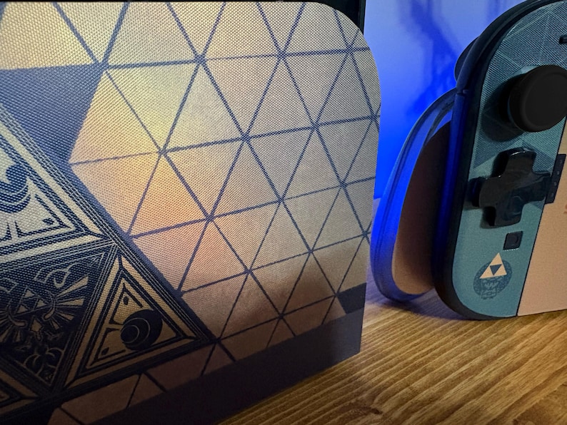 May include: A gaming console dock and a handheld controller featuring a blue and gold geometric triangle pattern. The controller displays a small gold triforce emblem and black buttons, while the dock shows a larger intricate triangular design.