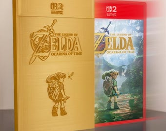 May include: A gold-colored game case cover featuring an embossed illustration of a character with a shield and sword, alongside the text The Legend of Zelda Ocarina of Time and Nintendo Switch logo, positioned next to a red game case with cover art.