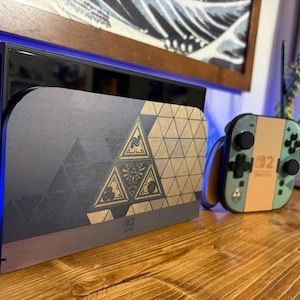 May include: A Nintendo Switch console dock and a matching Joy-Con controller featuring a gold and black geometric pattern with a central triangular emblem. The dock displays a dark grey finish with gold accents, while the controller is light green.