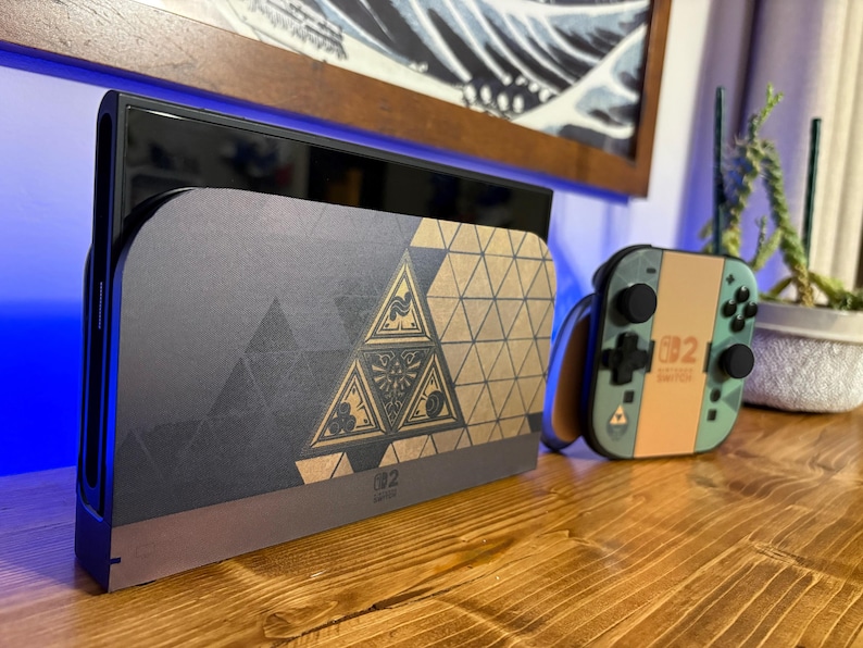 May include: A Nintendo Switch console dock and a matching Joy-Con controller featuring a gold and black geometric pattern with a central triangular emblem. The dock displays a dark grey finish with gold accents, while the controller is light green.