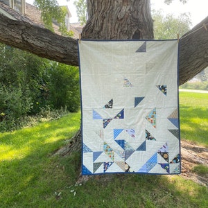 May include: A white quilt with a blue border, featuring a patchwork design of blue, white, and gray triangles. The quilt is hanging on a tree branch.
