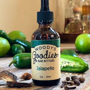 May include: A brown glass bottle of Woody's Goodies Bar Bitters, labeled "Jalapeño." The bottle has a black dropper top and is surrounded by green jalapeño peppers, limes, and spices on a wooden surface. The bottle contains 2 oz or 60ml.