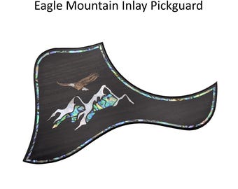 May include: A black guitar pickguard featuring an Eagle Mountain Inlay design. The pickguard displays an inlaid eagle flying above mountain peaks, accented with iridescent abalone shell borders and mountain details.