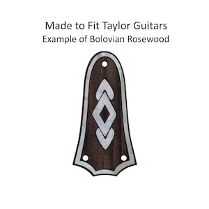 May include: A guitar headstock overlay made of Bolivian Rosewood. The dark brown wood is inlaid with white mother-of-pearl in a decorative pattern. The text "Made to Fit Taylor Guitars" and "Example of Bolivian Rosewood" is visible.