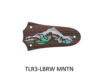 May include: A dark brown wooden truss rod cover featuring an iridescent abalone shell inlay shaped like a mountain range. The piece has three small circular mounting holes and the text TLR3-LBRW MNTN printed in black below the object.
