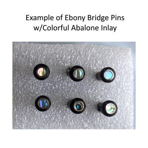 May include: Six ebony bridge pins with colourful abalone inlay are shown. Each pin has a dark, round head with a circular abalone inlay. The text "Example of Ebony Bridge Pins w/Colorful Abalone Inlay" is at the top.