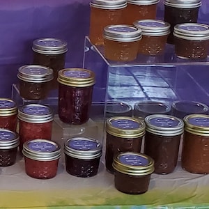 May include: A display of various jars of preserves, showcasing a range of colors from deep red to amber. Each jar has a silver lid and a purple label. The jars are arranged on clear acrylic risers and a green and purple surface.