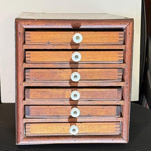Op de afbeelding: Een kleine houten kast met zeven laden. De kast is gemaakt van donkerbruin hout met lichtere ladefronten. Elke lade heeft een witte, ronde knop. De kast wordt waarschijnlijk gebruikt voor opslag.
