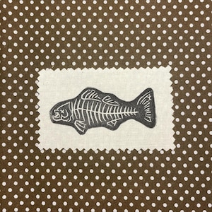 May include: A fabric square with a fish skeleton design. The fish skeleton is black and white, and the fabric is a light beige color. The background is brown with white polka dots.