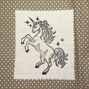 May include: A rectangular piece of off-white fabric with pinked edges featuring a black ink illustration of a rearing unicorn. The unicorn has a flowing mane and tail, a single horn, and is surrounded by four small four-pointed star shapes.
