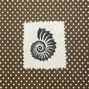 May include: A square fabric swatch with a black ammonite fossil design on a cream background. The swatch is set against a brown fabric with white polka dots, creating a contrasting pattern. The design is detailed and artistic.