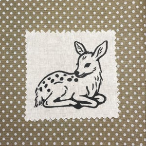 May include: A square fabric patch with pinked edges featuring a black ink illustration of a resting fawn. The patch is centered on a tan background fabric that is covered in a repeating pattern of small, evenly spaced white polka dots.