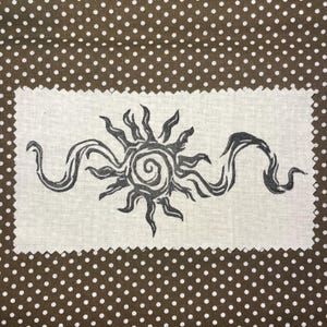 May include: A rectangular fabric patch with a gray sun design and flowing lines. The sun has a spiral center and spiky rays. The patch is on a brown background with white polka dots. The edges of the patch are unfinished.