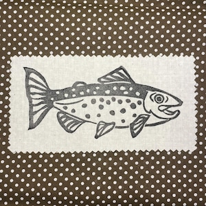 May include: A fabric square featuring a detailed illustration of a fish in shades of gray, with a dotted pattern. The fish is set against a white background with a scalloped edge, placed on a brown fabric with white polka dots. The design is suitable for home decor.