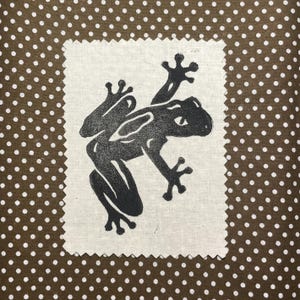 May include: A black frog illustration on a square, off-white fabric patch with a decorative, jagged edge. The patch is set against a brown background with white polka dots. The frog is depicted in a stylized, artistic manner.