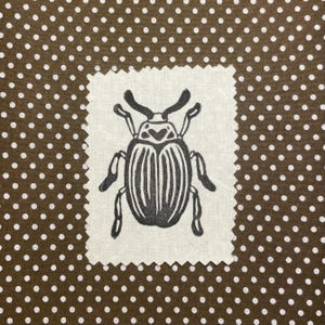 May include: A square fabric patch with a black beetle design. The beetle is centered on a white background with a jagged edge. The background is a brown fabric with white polka dots.
