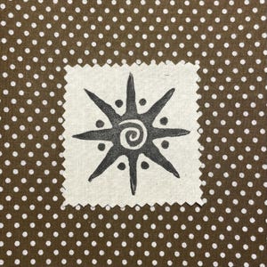 May include: A square, off-white fabric patch with a dark gray sunburst design featuring a central spiral. The patch is set against a brown background with white polka dots, creating a contrasting pattern.