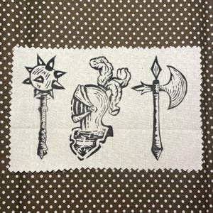 May include: A cream-colored fabric patch featuring three medieval weapon illustrations in black ink: a spiked mace, a knight's helmet, and a battle axe. The background is a brown fabric with white polka dots.