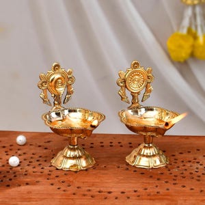 May include: Two gold metal diya oil lamps with intricate designs. Each lamp has a round base and a decorative top with a floral pattern. The lamps are lit with wicks and flames.