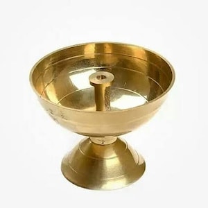 May include: A gold metal diya, a traditional Indian oil lamp, with a round bowl and a stand. The diya has a small hole in the center of the bowl for a wick.
