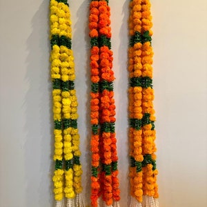 May include: Three garlands of artificial marigold flowers, each with alternating yellow, orange, and green sections. The garlands are hanging on a white wall.