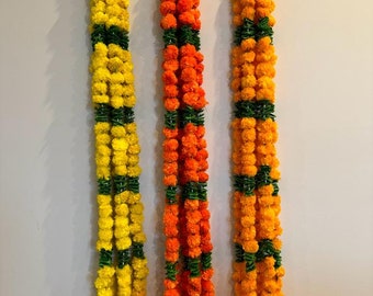 May include: Three garlands of artificial marigold flowers, each with alternating yellow, orange, and green sections. The garlands are hanging on a white wall.