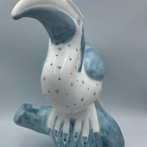 May include: A ceramic toucan ornament in white and blue. The bird is perched on a branch, with a white body speckled with blue star-shaped designs. The beak is white, and the wings and branch are blue.