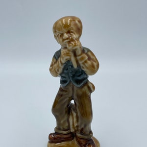 May include: A glazed ceramic figurine of a young boy in brown trousers, a dark vest, and brown shoes. The boy is holding a small object to his mouth. The figurine is standing on a small, oval base.
