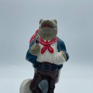 May include: A ceramic frog figurine, wearing a blue jacket, white shirt, and brown trousers, with a red necktie and a red bag over its shoulder. The frog stands on a grey base, with a white bandage on its arm.