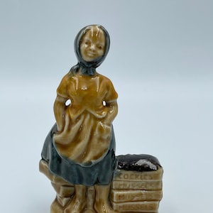 May include: A ceramic figurine of Molly Malone, a woman in a headscarf and apron, standing next to a stack of boxes labeled "Cockles" and "Mussels." The figurine is primarily brown, with accents of black and yellow. The base reads "Molly Malone."