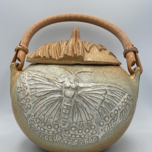 Puede incluir: Tetera de cerámica con un esmalte marrón claro y un diseño de mariposa tallada en la parte delantera. La tapa está rematada con un diseño tallado y texturizado. Tiene un asa de madera y es una pieza única de menaje de cocina.