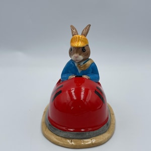 May include: A ceramic figurine of a brown rabbit wearing a blue jacket and yellow hat, driving a red car with black wheels.