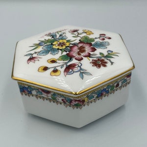 Könnte beinhalten: Eine weiße sechseckige Porzellan-Schmuckdose mit einem Blumendesign in Rot-, Gelb-, Blau- und Grüntönen. Der Deckel ist mit einem goldenen Rand und einem zarten Blumenmuster verziert.