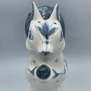 May include: A white ceramic squirrel ornament with blue floral and leaf patterns. The squirrel holds a blue sphere and has a fluffy tail. The figurine has a whimsical, hand-painted aesthetic, suitable for home decoration.
