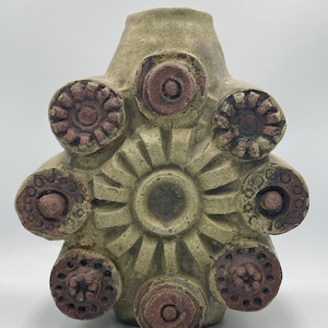 Puede incluir: Un jarrón de cerámica verde con una superficie texturizada y detalles marrones. El jarrón tiene una forma única con un círculo central grande y seis círculos más pequeños alrededor.