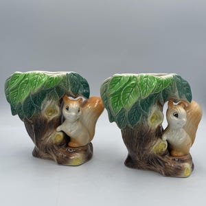 Puede incluir: Dos macetas de cerámica con forma de ardilla. Cada maceta presenta una ardilla posada en un tronco de árbol con hojas verdes. Las ardillas tienen pelaje marrón y colas naranjas. Las macetas están pintadas con colores marrón, verde y beige.