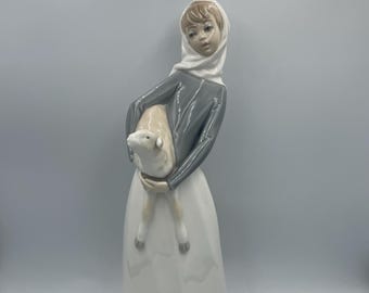May include: A porcelain figurine of a young person holding a lamb. The figure wears a gray top, a white headscarf, and a long white skirt with a gray hem. The lamb is white with gray legs.