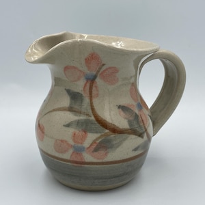 May include: A small, hand-painted ceramic pitcher with a floral design. The pitcher is cream-colored with pink and blue flowers and green leaves. The pitcher has a brown rim and a gray base.