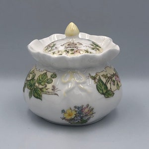 Op de afbeelding: Een witte keramische pot met een deksel, versierd met bloemen en botanische illustraties. Het deksel heeft het woord "Spring" en een gele knop. De pot heeft een gerimpelde rand en een strik.