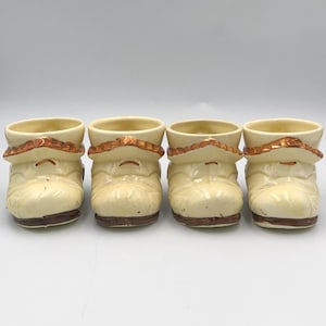 May include: Four ceramic boot-shaped shot glasses. The boots are off-white with brown soles and a decorative brown trim around the top. The shot glasses are arranged in a row against a white background.