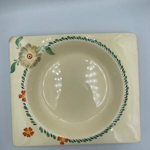 May include: Cream-colored, square-shaped ceramic bowl with a circular interior. The rim is decorated with a green and black vine pattern, and small orange flowers. A large flower with green leaves is on the corner.