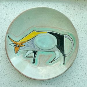 May include: A round ceramic plate featuring a stylised illustration of a bull. The bull is rendered in shades of yellow, brown, and black against a light, speckled off-white background with soft turquoise and pinkish-grey glaze accents.