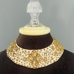 May include: A cream-colored, beaded collar necklace with gold floral and leaf designs. The collar is displayed on a mannequin. The necklace has a scalloped edge and is embellished with gold beads and sequins.