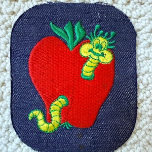 May include: Embroidered patch featuring a red apple with green leaves and two yellow and green worms. The patch is on a dark blue denim background with rounded corners. The design is a whimsical, cartoon-like illustration.