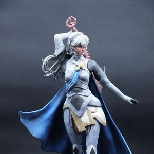 May include: A white and blue painted figurine of a female character with long white hair, wearing a blue cape and white armor. The figurine is posed with one arm raised and the other extended out to the side. The figurine is standing on a black base with a blue ring around the edge.