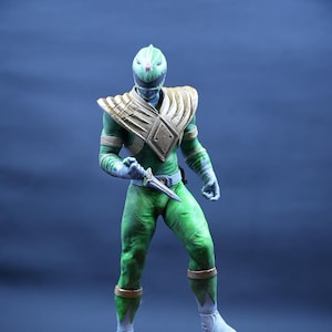 Può includere: Figurina del Power Ranger verde con una placca toracica dorata e un pugnale verde e argento. La figura è in piedi su una base rocciosa.
